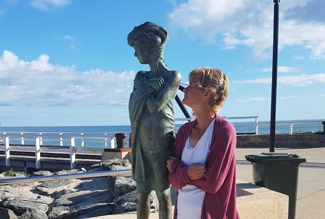 Sam is standing next to a statue of a young girl with her arms crossed in the same pose. Behind is a pier and the ocean.