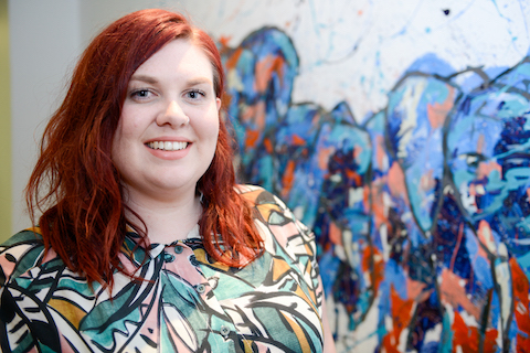 person with red hair and colourful shirt smiles at camera there is a wall with graffiti behind them