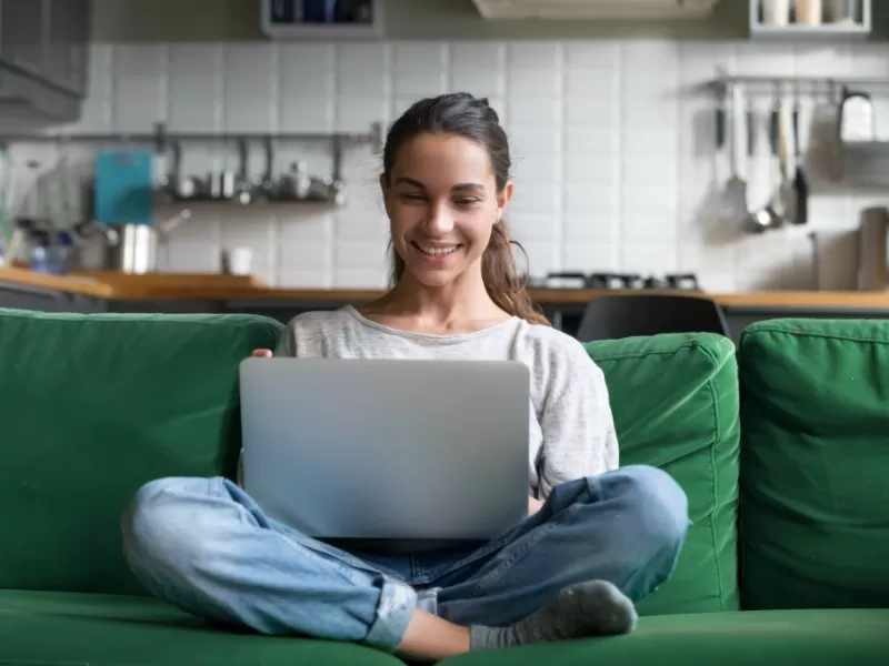 Young woman wearing blue jeans and a grey top, sitting cross legged on a green couch.